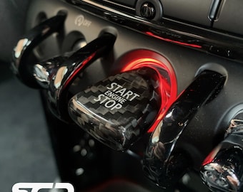 May include: Close-up of a black carbon fiber start engine stop button with a red illuminated ring around it. The button is in a car's dashboard.
