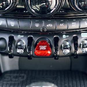 May include: A close-up of a car's dashboard with a red start engine stop button, black and silver controls, and a black and white carbon fibre pattern.