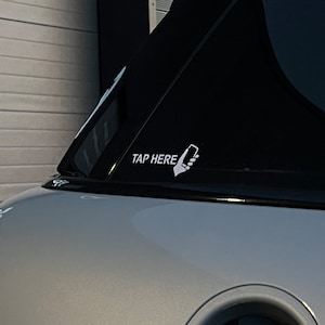 Op de afbeelding: Een witte sticker op een zwart autovenster met de tekst "TAP HERE" en een hand die een smartphone-icoon vasthoudt.