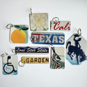 May include: Collection of keychains with various designs. Includes a square keychain with an orange, a rectangular keychain with the word "TEXAS", and a keychain with the word "Cali" in red.