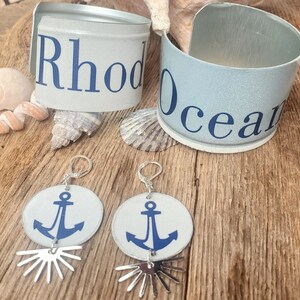 May include: Two pairs of earrings with silver hoops and white circular charms featuring blue anchor designs. The earrings are displayed on a wooden surface with two blue and white can bracelets that say "Rhode Island" and "Ocean".