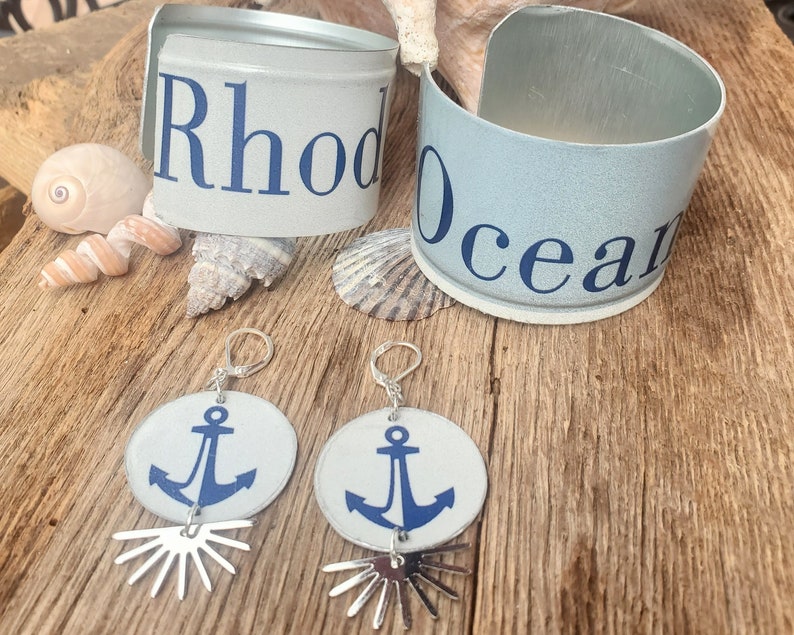 May include: Two pairs of earrings with silver hoops and white circular charms featuring blue anchor designs. The earrings are displayed on a wooden surface with two blue and white can bracelets that say "Rhode Island" and "Ocean".