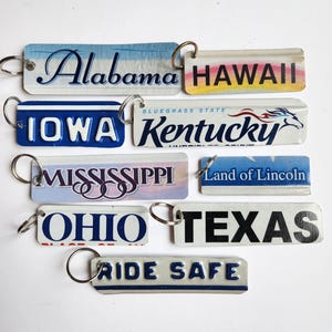 May include: A collection of rectangular keychains featuring state names and slogans. The keychains include Alabama, Hawaii, Iowa, Kentucky, Mississippi, Ohio, Texas, and "Ride Safe". Each keychain has a metal ring.
