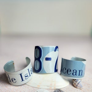 May include: Three blue and white metal bracelets with the words "de Isla", "8-2", and "ocean" on them. The bracelets are arranged on a white sandy surface with seashells and sea glass.