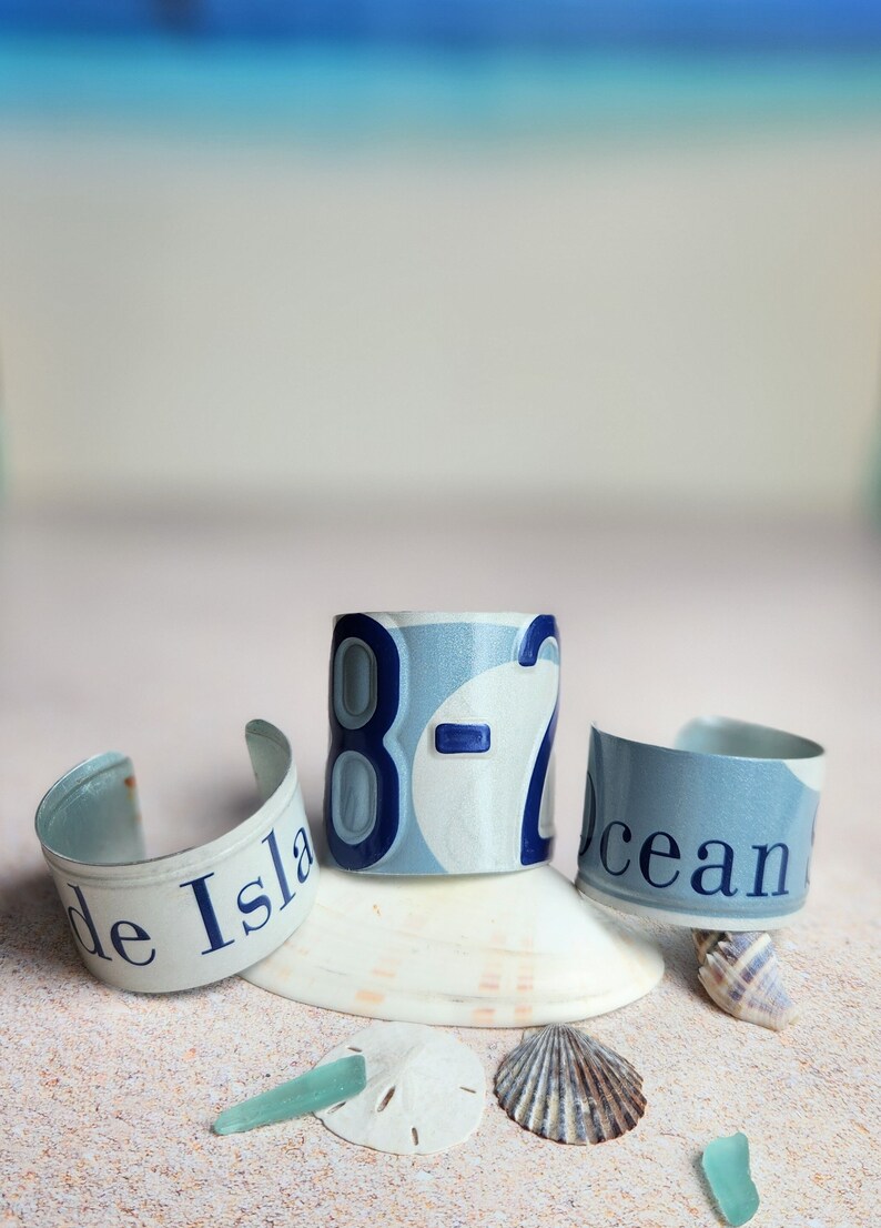 May include: Three blue and white metal bracelets with the words "de Isla", "8-2", and "ocean" on them. The bracelets are arranged on a white sandy surface with seashells and sea glass.