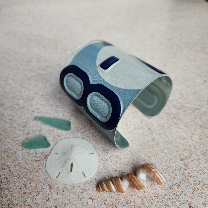 May include: A silver cuff bracelet with a blue and white design featuring the number "8". The bracelet is on a sandy surface with seashells and sea glass.