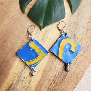 May include: A pair of earrings made from repurposed metal with blue and yellow paint. The earrings are shaped like a boomerang and a hook.
