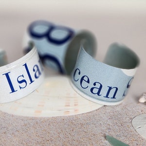 May include: Three white and blue fabric bracelets with the words "Blue Island" and "Ocean" printed on them. The bracelets are lying on a sandy surface with a seashell and a sand dollar.