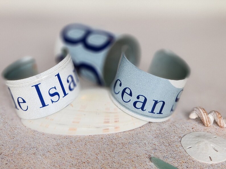 May include: Three white and blue fabric bracelets with the words "Blue Island" and "Ocean" printed on them. The bracelets are lying on a sandy surface with a seashell and a sand dollar.