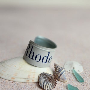May include: A silver metal cuff bracelet with the word "Rhode" printed in blue on a white background. The bracelet is resting on a seashell and surrounded by other seashells and sea glass on a sandy beach.