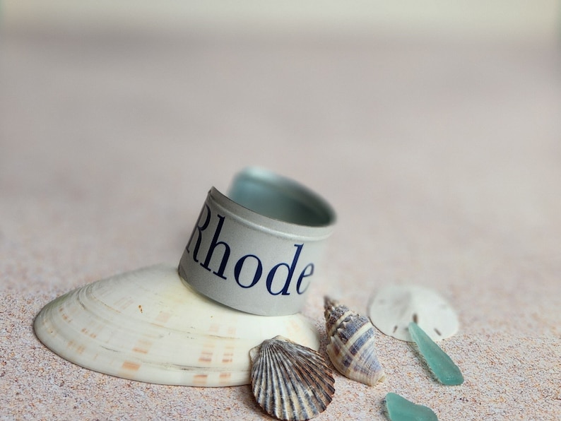 May include: A silver metal cuff bracelet with the word "Rhode" printed in blue on a white background. The bracelet is resting on a seashell and surrounded by other seashells and sea glass on a sandy beach.