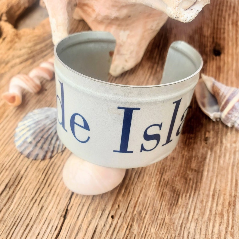 May include: A white metal cuff bracelet with the words "de Isla" printed in blue.