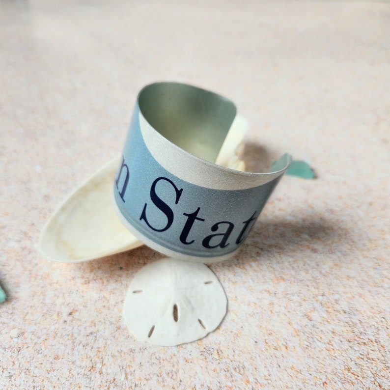 May include: A blue and white metal cuff bracelet with the words "In Stat" printed on it. The bracelet is lying on a white surface with a seashell.