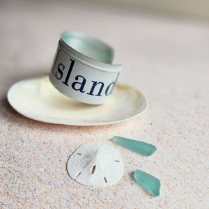 May include: A white seashell with a blue and white metal band that says "Island" on it. There are two small pieces of turquoise sea glass on a sandy surface.