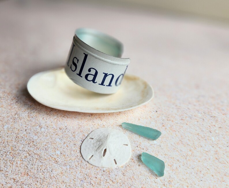 May include: A white seashell with a blue and white metal band that says "Island" on it. There are two small pieces of turquoise sea glass on a sandy surface.