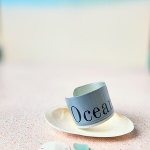 May include: A light blue and white bracelet with the word "Ocean" printed on it. The bracelet is sitting on a white seashell on a sandy beach with two pieces of sea glass.