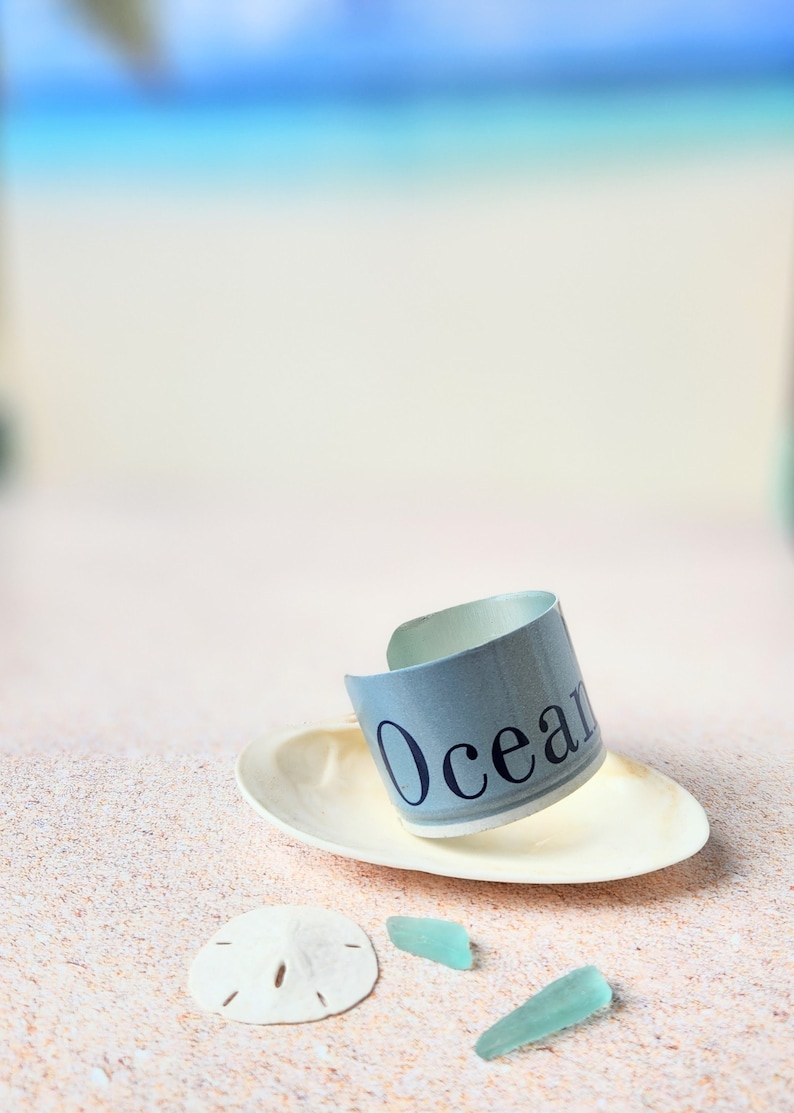 May include: A light blue and white bracelet with the word "Ocean" printed on it. The bracelet is sitting on a white seashell on a sandy beach with two pieces of sea glass.