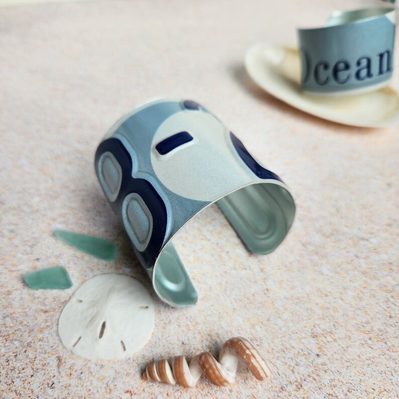 May include: A silver metal cuff bracelet with a blue and white abstract design. The bracelet has the word "ocean" printed on it.