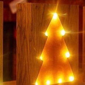 May include: A wooden Christmas tree-shaped light. The tree is cut out of a rectangular block of wood, with warm yellow lights illuminating the tree shape. The wood has a natural grain.