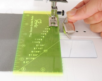 May include: A green plastic seam guide ruler with markings in inches and fractions of an inch. The ruler is labeled "The Seam Guide Ruler" and has a small logo that says "Madan Sew". The ruler is being used to guide fabric through a sewing machine.