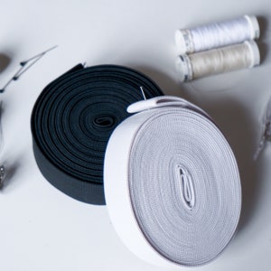 May include: Two rolls of elastic ribbon, one black and one white, are the focal point of this image. Sewing pins and spools of thread are also visible. The elastic is tightly wound, showing its texture. This image is ideal for sewing and craft supplies.
