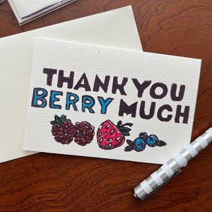 Puede incluir: Una tarjeta blanca con el texto "THANK YOU BERRY MUCH" en letras negras y azules, con ilustraciones de bayas. Un bolígrafo plateado y blanco descansa sobre la superficie de madera junto a la tarjeta y un sobre blanco.