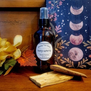 Op de afbeelding: Een bruine glazen fles met een spuitmond, gevuld met Palo Santo etherische olie. De fles heeft een wit etiket met de tekst "Smudge Your Space Palo Santo Witchful Intentions Net Vol. 4 oz". Op de voorgrond ligt een stuk Palo Santo hout.