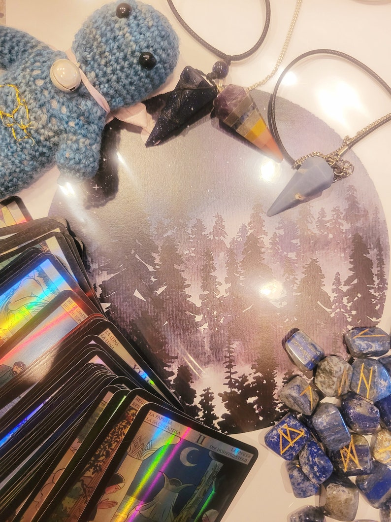 Pu&ograve; includere: Un peluche blu lavorato a uncinetto, un mazzo di carte dei tarocchi, due pendolari di cristallo e una collezione di pietre di lapislazuli blu con rune incise. Lo sfondo &egrave; un dipinto ad acquerello di una foresta illuminata dalla luna.