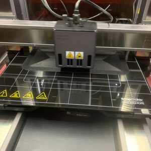 May include: A black 3D printer with a glass build plate that has a grid pattern and yellow warning signs. The text "snapmaker glass build plate" is printed on the plate.
