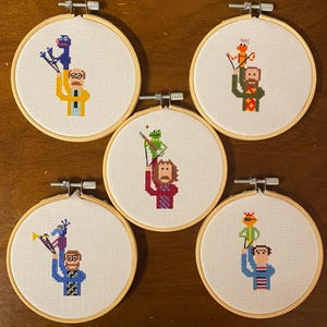 May include: Five circular wooden embroidery hoops containing cross-stitch patterns of various Muppet characters being held by their respective human puppeteers. Each hoop features a white fabric background with colorful pixelated character designs.
