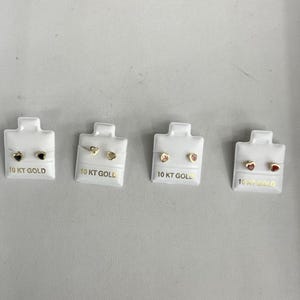 May include: Four pairs of heart-shaped earrings on display cards. The earrings are made of 10 karat gold and have different colored stones: black, white, pink, and red.