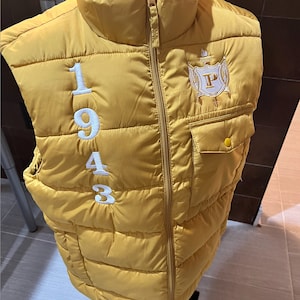 May include: A mustard yellow puffer vest with a white zipper and a white embroidered crest on the right side. The crest features a white letter "P" inside a shield with a torch above it. The vest has a white embroidered number "1943" on the left side.