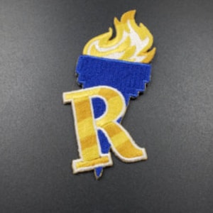 May include: An embroidered patch with a blue torch and a yellow 'R' with stripes. The torch has yellow flames.