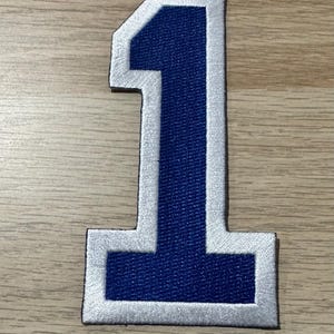 Könnte beinhalten: Gestickter Aufnäher in Form der Zahl eins. Die Zahl ist marineblau mit silberner Umrandung. Geeignet für Sportbekleidung oder Bastelprojekte.