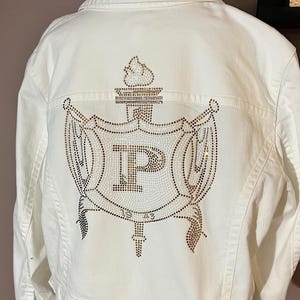 May include: White denim jacket with a large rhinestone design on the back. The design features a crown, a sword, and the letter "P" surrounded by a wreath.