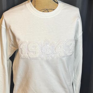 May include: White crew neck sweatshirt with the year "1943" embroidered in a textured, off-white thread. The design includes a crest with the letter "P" in the center. The sweatshirt is displayed on a mannequin.