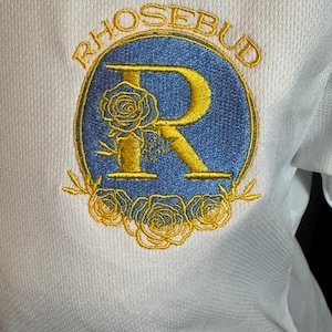May include: White athletic shirt with an embroidered emblem. The emblem features the word "RHOSEBUD" in gold lettering above a blue oval with a gold "R" and rose details. The shirt has a textured, breathable fabric.