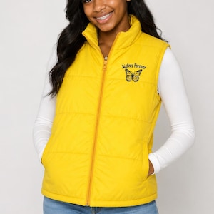May include: A bright yellow puffer vest featuring a stand-up collar and a full-length front zipper. The left chest area displays a black embroidered butterfly graphic positioned above the cursive black text that reads Sisters Forever.