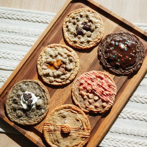 Könnte beinhalten: Sechs runde Kekse auf einem rechteckigen Holztablett. Die Kekse sind mit verschiedenen Zutaten belegt, darunter dunkle Schokoladenstückchen, Schokoladenguss mit bunten Streuseln, weiße Creme mit Keksbröseln, rote Glasurstreifen und orangefarbene Linien.