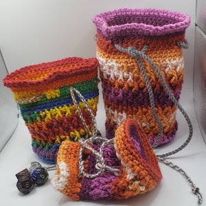 Puede incluir: Dos bolsas de dados de crochet en colores arcoíris con un cordón de ajuste gris. La bolsa más pequeña está tumbada de lado, y la más grande está de pie. Hay tres dados en la mesa.