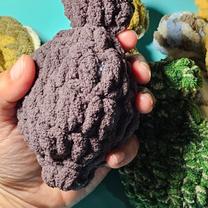 May include: A close-up of a hand holding a purple crocheted stuffed toy. The toy is shaped like a bird and has a textured surface. There are other crocheted toys in the background, including a green one and a blue and white one.