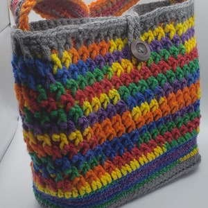 May include: A rainbow crochet tote bag with a gray strap and a brown button. The bag is made with a variety of colorful yarns.