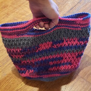 May include: A crocheted tote bag with a pink, purple and grey striped pattern. The bag has a single handle.