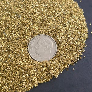 May include: A pile of gold glitter, with a US dime coin for scale. The glitter is fine and sparkly, and would be perfect for crafts or decorations.