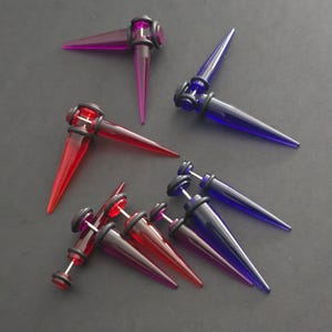 Puede incluir: Un juego de seis pares de dilatadores de orejas de acrílico en rojo, azul y morado. Cada par presenta un diseño acampanado y puntiagudo con una arandela negra.