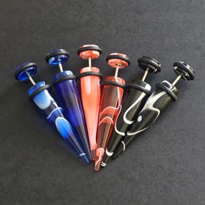 Puede incluir: Conjunto de seis pendientes de cono de acrílico con una variedad de colores y patrones, incluyendo blanco y negro, rojo y blanco, azul y blanco, y azul sólido. Los conos son todos puntiagudos en el extremo y tienen una junta tórica de goma negra en la parte superior.