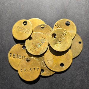 May include: A collection of antique brass tags, each round with a hole at the top. The tags have stamped numbers in a variety of fonts. The tags are a warm, aged gold color, with some showing signs of wear.