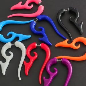 Puede incluir: Una colección de pendientes coloridos en forma de llama en varios tonos, incluyendo rosa, azul, negro, naranja, blanco, rojo y morado. Cada pendiente tiene un poste para perforar. Los pendientes se muestran sobre una superficie oscura.
