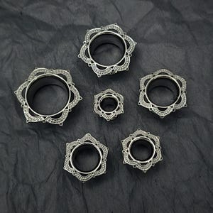 Peut inclure: Plusieurs écarteurs d'oreille argentés en forme de fleur. Les écarteurs présentent un design ajouré avec un trou central. Les bijoux sont disposés sur une surface sombre et texturée, mettant en valeur les détails complexes de chaque pièce.