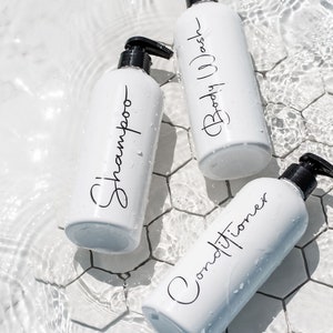 May include: Three white bottles with black pump tops and labels. The labels read "Shampoo", "Conditioner", and "Body Wash". The bottles are sitting on a white tiled surface with water droplets.